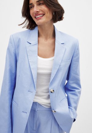 Light blue linen blazer featuring a notched lapel, two front pockets, and a single button closure. Worn over a white tank top.