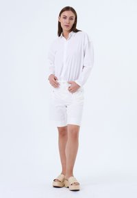 Witte lange mouw knoopsluiting shirt, gecombineerd met witte korte broek met omslagen en gevlochten platform sandalen. Soepele stof en lichtgewicht ontwerp.