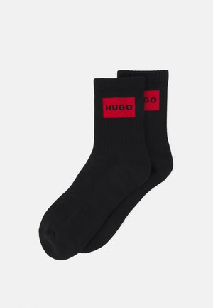 Schwarze Socken mit gerippter Textur, die ein rotes rechteckiges Logo mit dem Text "HUGO" auf der Oberseite gut sichtbar zeigen.