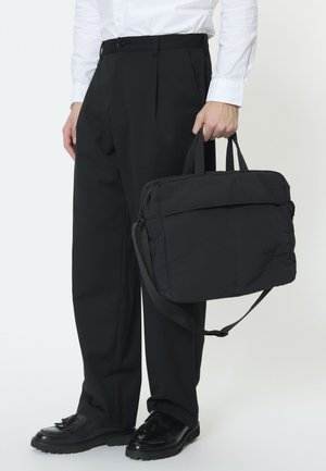 Man draagt zwarte broek en zwarte schoenen, houdt een zwarte aktetas vast aan de handvatten met een schouderriem.