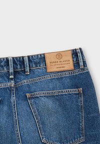 Jeans en denim ; tissu bleu foncé ; étiquette en cuir avec logo embossé ; deux poches arrière ; surpiqûres contrastées ; design décontracté.