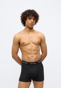 Boxeri scurți negri din bumbac, cu un fit strâns, având un elastic neted inscripționat "Calvin Klein". Modelul are o constituție musculară și păr creț.