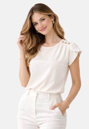 Kobieta o długich falowanych włosach nosi białą bluzkę z krótkimi rękawami z złotymi guzikami na ramieniu oraz białe spodnie z wysokim stanem, uśmiechając się.