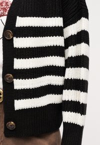 Cardigan tricoté à rayures noires et blanches avec de gros boutons marron, porté sur un haut à fleurs rose et blanc et un pantalon marron.