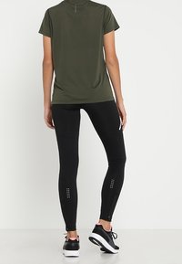Svarta leggings med reflexdetaljer och sidoknappar, tillsammans med en mörkgrön t-shirt med kort ärm och en diskret detalj på baksidan.