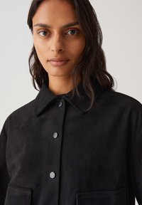 Giacca in suede nera con colletto a punta, due tasche frontali e bottoni a pressione. Texture liscia e vestibilità leggermente oversize.
