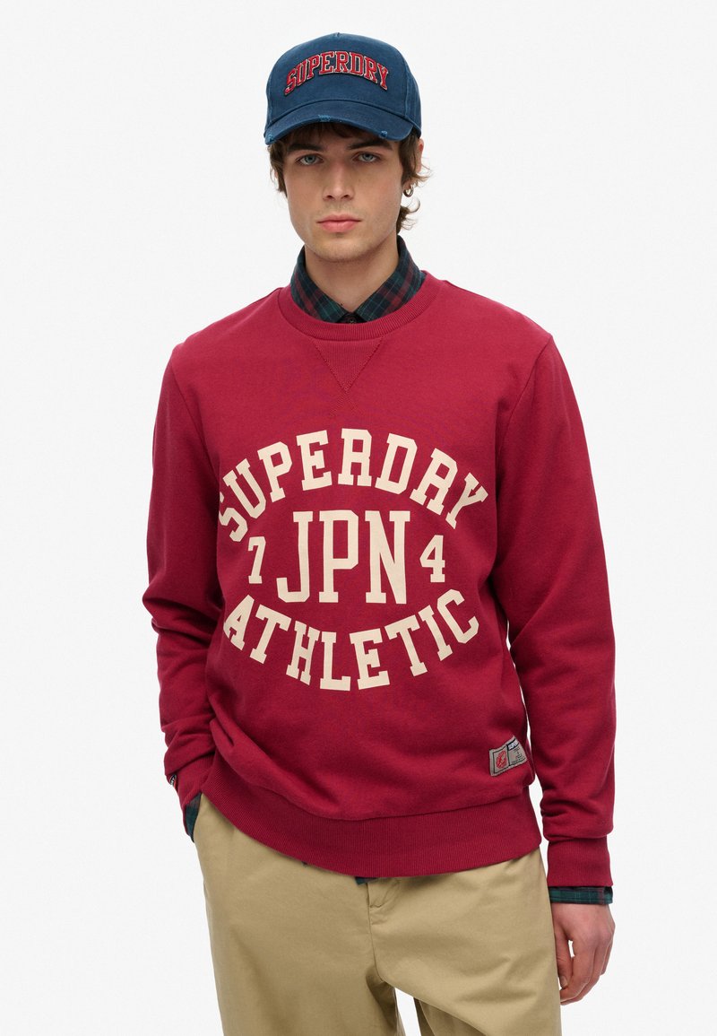 Czerwona bluza z dużym białym nadrukiem "SUPERDRY JPN 4 ATHLETIC", okrągły dekolt, ściągacze przy rękawach i na dole. Noszona z granatową czapką i koszulą w kratę.