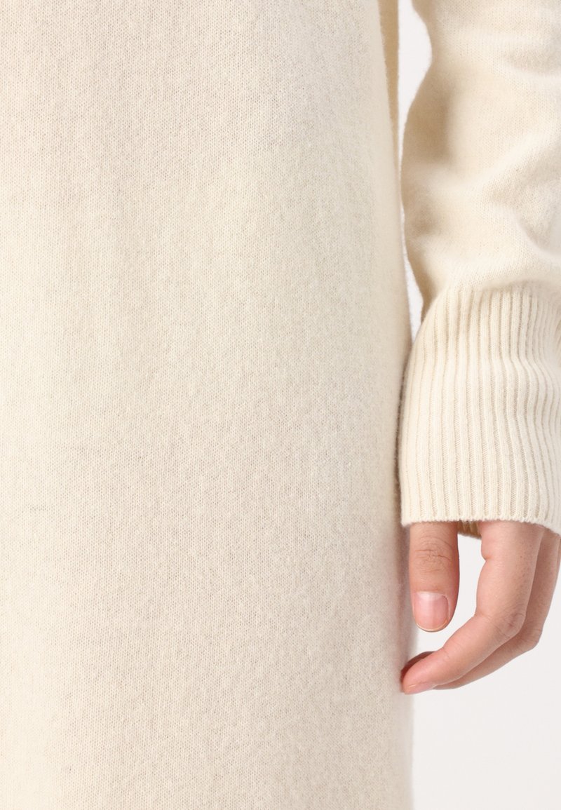Kremowy sweter z dzianiny z prążkowanymi rękawami i miękką teksturą, z dopasowanym krojem i lekkim opończeniem. Ręka opiera się na tkaninie.