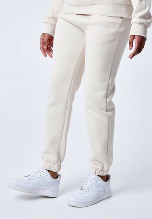 Pantalons de survêtement couleur crème avec des poignets élastiques et des poches latérales, présentant un détail de logo brodé sur la hanche, portés avec des baskets blanches.