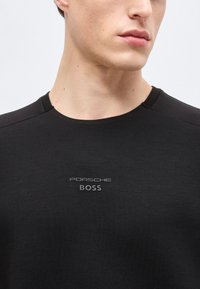 Jeune homme portant un t-shirt noir à col rond avec le logo "PORSCHE BOSS" centré sur la poitrine, légèrement tourné vers la droite.