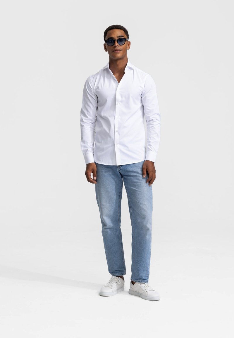 Witte longsleeve overhemd met knopen, lichtblauwe jeans en witte sneakers. Ronde zonnebril en een ontspannen pasvorm benadrukken de casual stijl.