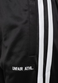 Crne atletske kratke hlače s bijelim vertikalnim prugama. Imaju teksturiranu tkaninu i sašiveni natpis "UNFAIR ATHL." sa strane.