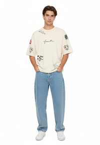 Overdimensioneret cremefarvet t-shirt med grafiske designs og tekst, kombineret med lyseblå straight-leg jeans og hvide sneakers.