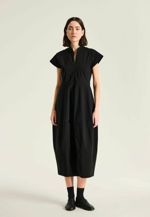 Femme debout portant une robe midi noire à manches courtes, une fermeture éclair frontale, des chaussettes noires et des chaussures noires sur un fond clair uni.