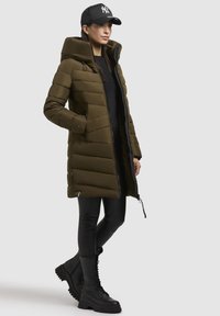 Manteau rembourré vert olive avec capuche, fermeture éclair à l'avant et poches latérales. Associez-le à des leggings noirs et des bottes de combat noires à lacets pour un look cohérent.