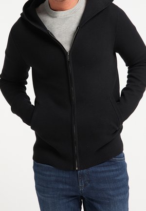 Homme portant un sweat à capuche noir zippé sur un t-shirt gris clair et un jean bleu, les mains dans les poches du sweat.