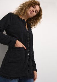 Svart, strukturerad cardigan med volangkanter, knäppning fram och en framficka. Materialet verkar mjukt och lätt skrynkligt.