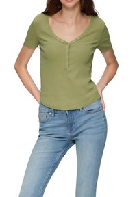 Groene ribbel top met korte mouwen, een V-hals en een knoopsluiting, gecombineerd met lichtblauwe jeans. De stof lijkt glad en strak te zitten.