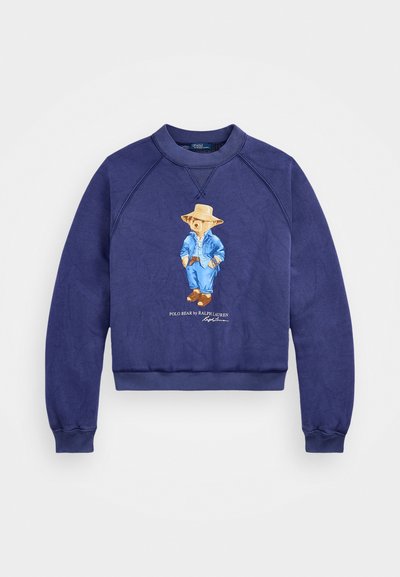 Σκούφος ναυτικού μπλε με κυκλικό λαιμό και ραγλάν μανίκια. Περιέχει μια απεικόνιση αρκούδας ντυμένη με μπλε τζιν και το κείμενο "POLO BEAR by RALPH LAUREN."