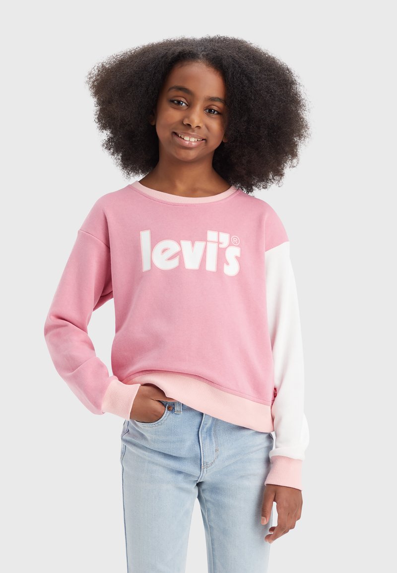 Fille souriante avec des cheveux afros portant un sweat-shirt Levi's rose et blanc et un jean bleu clair, se tenant avec une main dans sa poche.