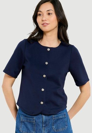 Vrouw draagt een navy blauw kortemouwen top met decoratieve zilveren knopen, gecombineerd met blauwe spijkerbroek.