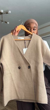 Cardigan in maglina beige con scollo a V, design doppiopetto, caratterizzato da due bottoni scuri. Tessuto morbido e vestibilità rilassata. Etichetta di sospensione visibile.