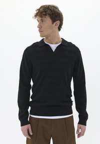 Suéter de punto a rayas negro con escote en V y cuello, combinado con una camisa blanca debajo, acompañado de pantalones marrones texturizados.