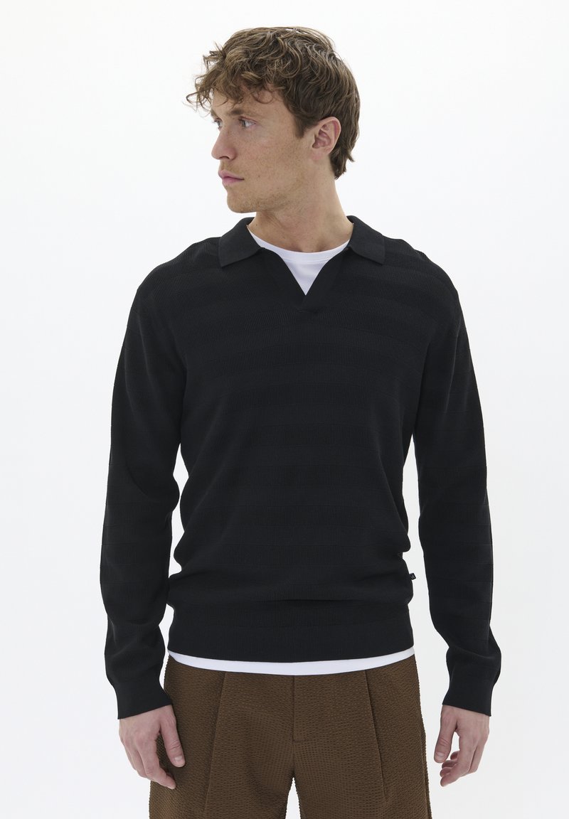 Suéter de punto a rayas negro con escote en V y cuello, combinado con una camisa blanca debajo, acompañado de pantalones marrones texturizados.