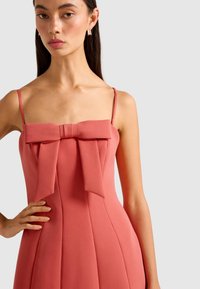 Frau, die ein ärmelloses pinkes Kleid mit dünnen Trägern und einer großen Schleifendetail an der Brust trägt, mit der Hand auf der Hüfte, vor einem schlichten hellen Hintergrund.