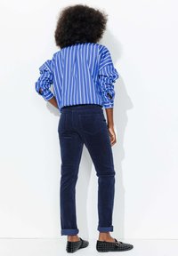 Chemise rayée bleu et blanc, pantalon bleu marine et mocassins noirs à clous. La chemise a des manches retroussées ; le pantalon est ajusté avec des poches au dos.
