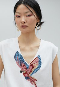White cotton top with a deep V-neck featuring colorful embroidered bird design in blue, pink, red, and gold threads.