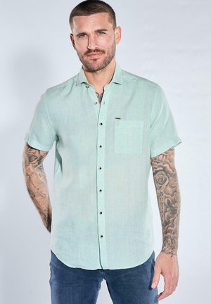 Mann mit kurzen Haaren und tätowierten Armen, der ein hellgrünes kurzärmeliges Hemd mit Knöpfen und dunkelblaue Jeans trägt, vor weißem Hintergrund stehend.