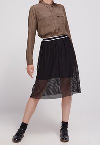 Blusa marrone a maniche lunghe con due tasche sul petto, abbinata a una gonna midi in rete a pois neri e scarpe nere con lacci. Fascetta bianca in evidenza.