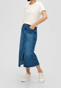 Džínová sukně střední délky s předním rozparkem a bočními kapsami, doplněná jednoduchým bílým tričkem a béžovými teniskami.