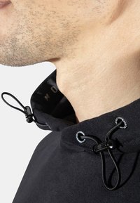 Sudadera negra con cuello ajustable. Presenta un herraje negro y una tela texturizada. La imagen en primer plano muestra el área del cuello y los detalles del collar.
