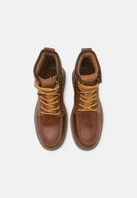Botas de cuero marrón con cordones amarillos, cuello acolchado y costuras en contraste. El diseño presenta una punta redondeada y una suela de goma resistente.