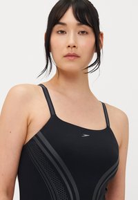 Maillot de bain noir avec des bretelles fines, présentant des accents gris courbes et un motif en nid d'abeille sur les côtés. Texture lisse et ajustée.