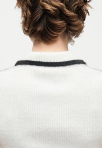 Κοντά καστανά κυματιστά μαλλιά που φαίνονται από πίσω σε ένα άτομο που φοράει λευκό πουλόβερ με υφή και μαύρη ρίγα κοντά στον γιακά.