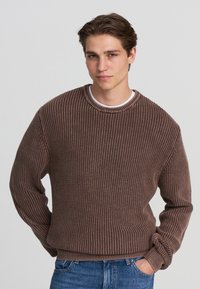 Maglione marrone a coste con scollatura rotonda e maniche lunghe, abbinato a jeans azzurri chiari. Tessuto testurizzato con vestibilità morbida.
