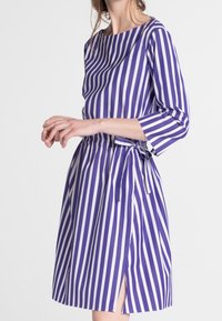 Fialové a bílé pruhované šaty s třičtvrtinovými rukávy, lodičkovým výstřihem, elastickým pasem a bočním rozparkem. Obsahují detail s opaskem na zavazování.