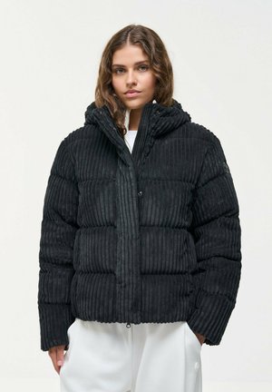 Une femme porte une doudoune noire à capuche, à la texture côtelée, par-dessus des vêtements blancs, sur un fond clair uni.