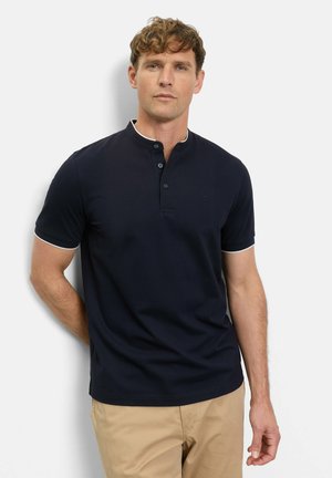 Man draagt een marineblauw henley-shirt met korte mouwen en beige broek, staand tegen een witte muur met zijn handen achter zijn rug.