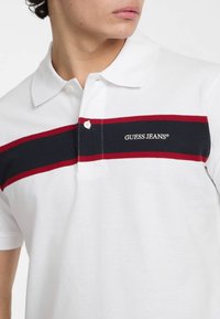 Camisa polo blanca con un diseño de rayas rojas y marinas, logotipo bordado de "GUESS JEANS", mangas cortas y cuello clásico.