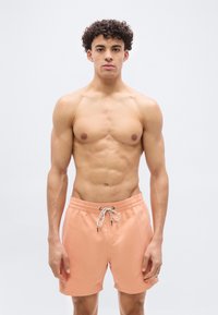 Νέος άνδρας με σγουρά μαλλιά, φοράει ροδακινί μαγιό με λευκό κορδόνι, στέκεται μπροστά σε απλό ανοιχτό φόντο, κοιτάζοντας προς τα εμπρός.