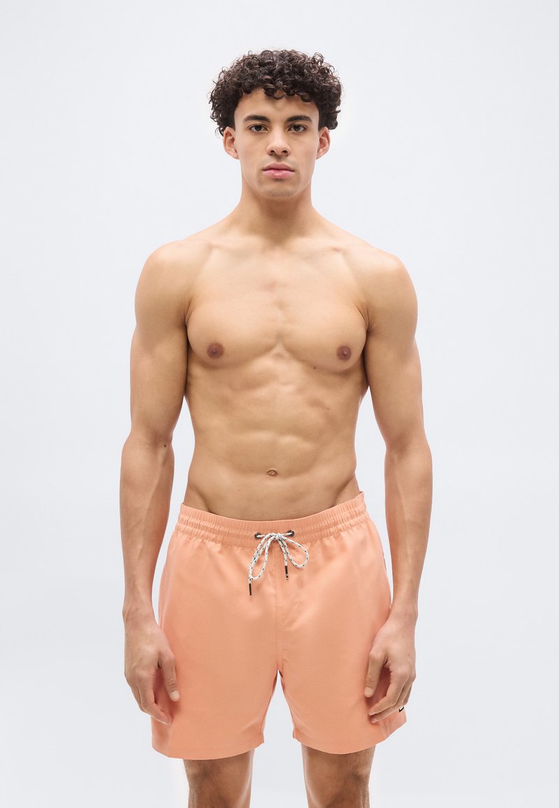 Νέος άνδρας με σγουρά μαλλιά, φοράει ροδακινί μαγιό με λευκό κορδόνι, στέκεται μπροστά σε απλό ανοιχτό φόντο, κοιτάζοντας προς τα εμπρός.