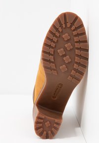 Semelle extérieure en caoutchouc d'une botte de travail beige, inclinée contre un coin blanc, montrant des rainures profondes et des motifs de grip rectangulaires.