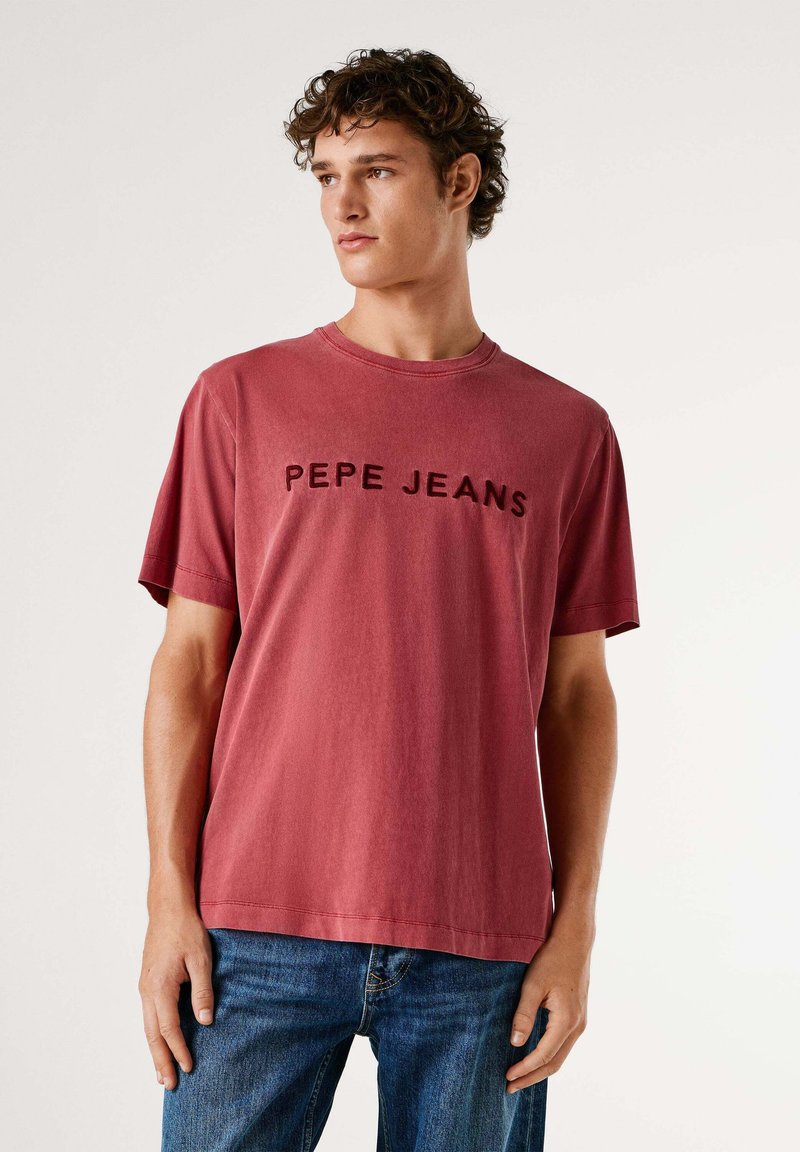 Camiseta de algodón roja con el texto "PEPE JEANS" en negro, mangas cortas, cuello redondo, ajuste relajado, combinada con vaqueros azules de mezclilla.