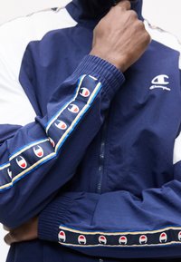 Persona con chaqueta azul marino de Champion con franjas con el logo a lo largo de las mangas y cremallera al frente, mano cerca de la cara, brazos cruzados.