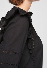 Schwarze Bluse mit Rüschkragen und -ärmeln, die offene Spitzenverzierungen entlang der Ärmel und des Körpers aufweist. Weicher Stoff.