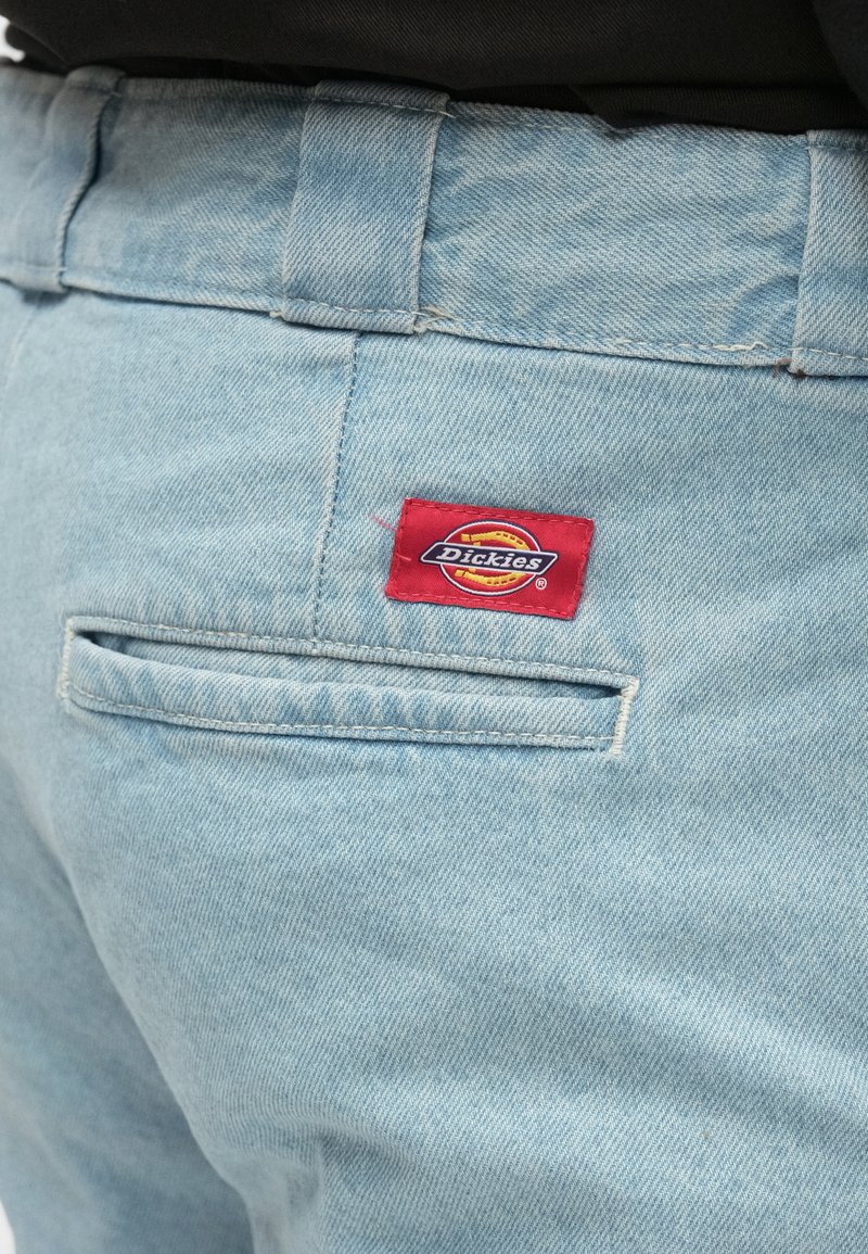 Pantalon en denim bleu clair avec une poche arrière et une étiquette rouge portant le logo "Dickies" en jaune et noir. Détail de couture visible.